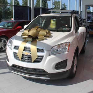 May include: A white Chevrolet Trax SUV with a gold bow on the hood. The car is parked in a showroom with a red car in the background.
