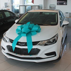 May include: A white Chevrolet sedan with a large turquoise bow on the hood. The car is in a showroom with a tiled floor.