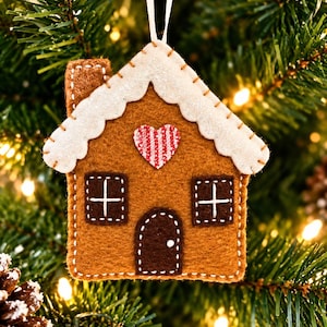 May include: A brown felt gingerbread house ornament featuring a white glittery roof, a red and white striped heart, two square windows with white cross stitching, and a dark brown arched door with a single white dot, suspended by a white ribbon.