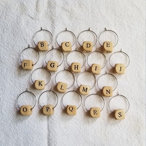 May include: Set of 19 wooden wine glass charms with metal rings. Each charm features a letter of the alphabet, from A to S. The charms are beige and have black lettering.