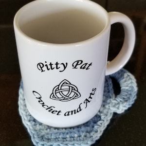 May include: White ceramic mug with the text "Pitty Pat Crochet and Arts" and a black Celtic knot design. The mug is sitting on a blue crocheted coaster.