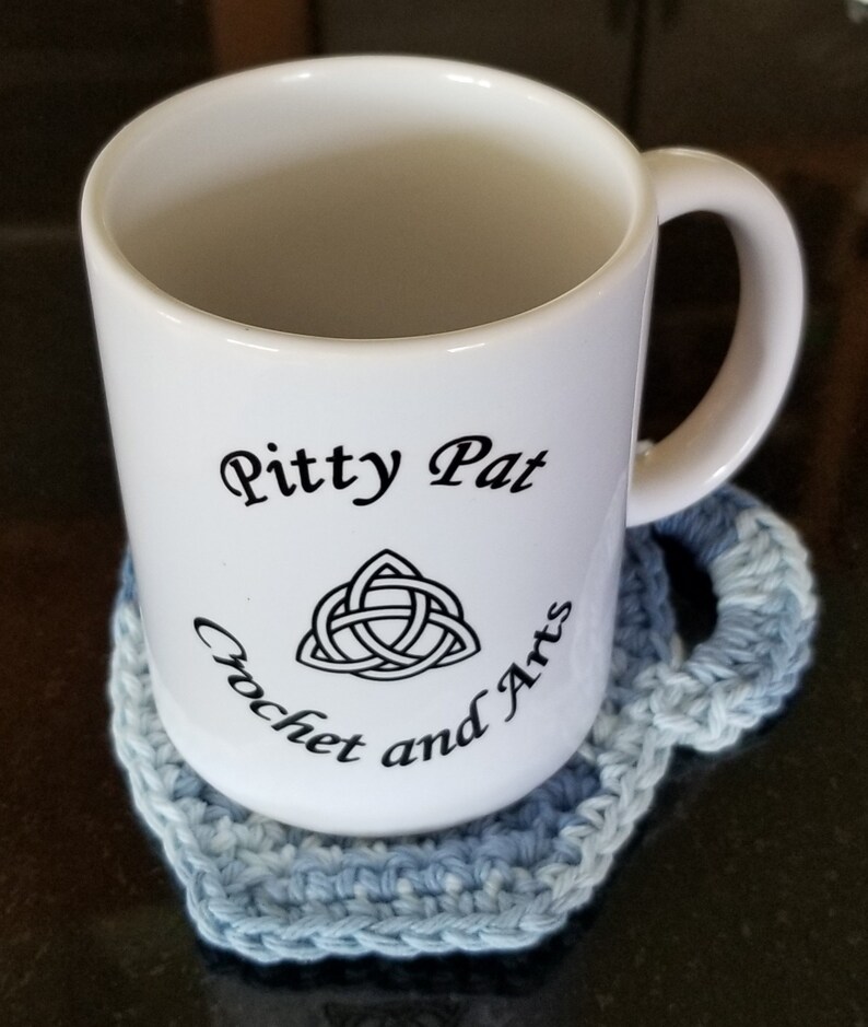 May include: White ceramic mug with the text "Pitty Pat Crochet and Arts" and a black Celtic knot design. The mug is sitting on a blue crocheted coaster.