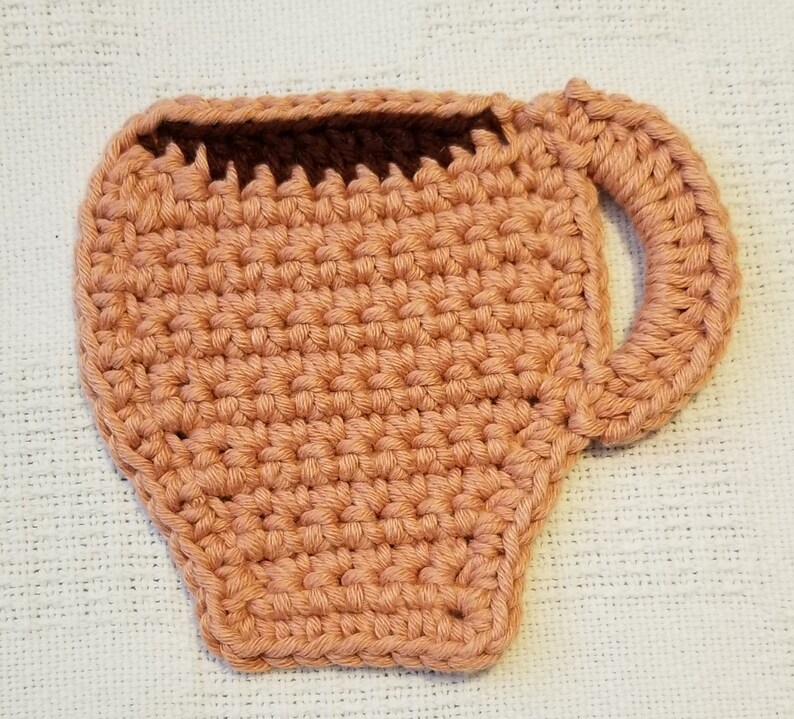 May include: A crocheted coffee cup coaster in a peach colour with a brown crocheted rim.
