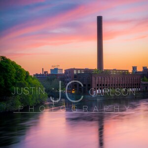 May include: A brick building with a tall smokestack against a colorful sunset sky. The building is reflected in the still water of a river.