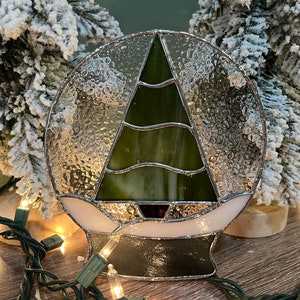 May include: A stained glass Christmas tree bauble featuring a green tree and white snow design. The bauble is set against a snowy backdrop.