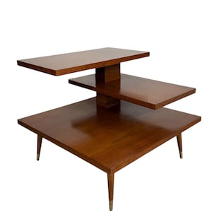 May include: A mid-century modern tiered side table with three rectangular surfaces. The table is made of wood with a warm brown finish. The legs are tapered with brass-colored tips. The top tier is approximately 12 inches long, the middle tier is 24 inches long, and the bottom tier is 30 inches long.