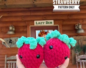 Puede incluir: Dos juguetes de peluche de fresa tejidos a crochet sin costuras, con ojos negros y hojas verdes. Los juguetes se sostienen en una mano frente a una cabaña de madera con un letrero que dice "Lazy Den".