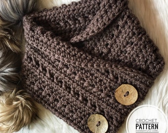 May include: Brown crocheted cowl with two wooden buttons. The cowl is folded in half and the buttons are visible on the folded edge. The cowl is on a white fuzzy surface.