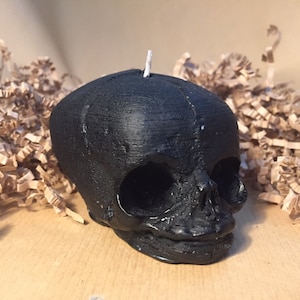 May include: A black candle shaped like a human skull. The candle has a detailed skull design with eye sockets, a nose, and a mouth.