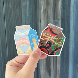 May include: Two cartoon-style beverage cartons. The blue carton says "Holy Water" with a small angel wearing a bow tie on the front. The brown carton says "Demonic Espresso" with a black snake wrapped around it and the text "Unleash Your Hellish Energy".