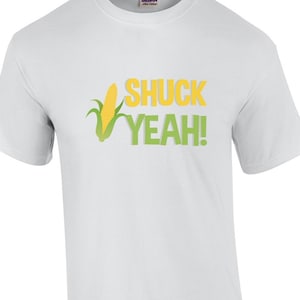 Può includere: Maglietta bianca con una grafica di mais verde e giallo e il testo "Shuck Yeah!"