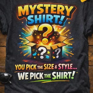 Può includere: T-shirt grigio scuro con grafica colorata. Il design presenta le parole "MYSTERY SHIRT!" sopra una scatola aperta con punti interrogativi e il testo "YOU PICK THE SIZE & STYLE... WE PICK THE SHIRT!"