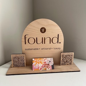 Op de afbeelding: Houten displaystandaard met het woord "found" en de tekst "sustainable • artisanal • luxury". Inclusief een visitekaartje en QR-codeblokken voor Google Reviews en Instagram. De standaard is gemaakt van lichtgekleurd hout.