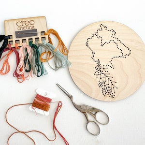 May include: A craft kit with a wooden base featuring a dotted outline of a landmass. The kit includes colored threads, a wooden needle holder with the text "creo crafting", and a pair of decorative scissors. The threads are in various colors.