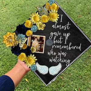May include: A black graduation cap decorated with yellow and blue paper flowers, a butterfly, a photo, and white angel wings. The cap has white text that reads "I almost gave up but I remembered who was watching."