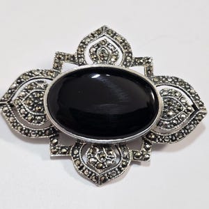 Op de afbeelding: Een zilveren broche met een zwarte ovale steen in een bloemmotief. De broche is gemaakt met marcasietstenen.