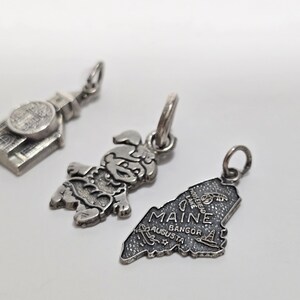 May include: Three silver charm pendants. One is a building, one is a cartoon character, and one is the outline of the state of Maine with the text "Maine" and the cities "Augusta, Bangor".