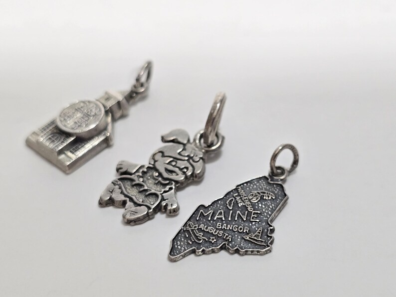 May include: Three silver charm pendants. One is a building, one is a cartoon character, and one is the outline of the state of Maine with the text "Maine" and the cities "Augusta, Bangor".
