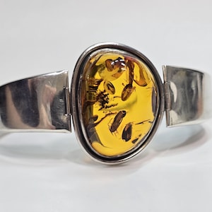 Könnte beinhalten: Ein silbernes Armband mit einem großen, polierten, tropfenförmigen Bernstein als zentralem Blickfang. Der Bernstein ist durchscheinend goldgelb mit sichtbaren dunklen organischen Einschlüssen und in einen glatten, polierten Silberrahmen gefasst.