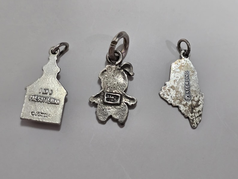 May include: Three silver charm pendants. One is a church with the text "1873 Presbyterian", another is a small person with a backpack, and the last is a state outline with the text "Sterling".