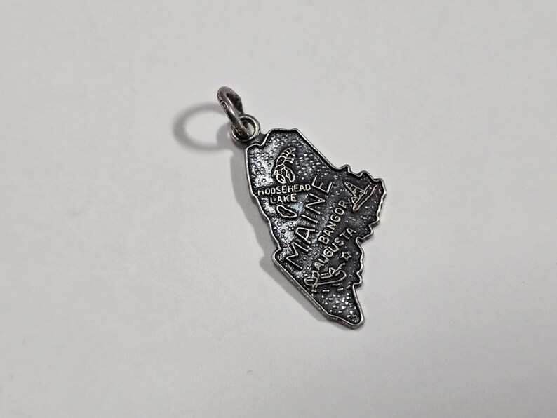 May include: Silver charm shaped like the state of Maine with the word "Maine" and the names of cities including "Moosehead Lake", "Bangor", and "Augusta" written on it.