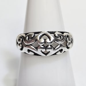 May include: A silver-colored ring with an openwork design featuring swirling, symmetrical patterns. The ring is displayed on a white surface, showcasing its intricate details and polished finish. The ring's design is reminiscent of floral or ornamental motifs.