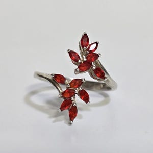 May include: A silver ring featuring a floral design with red gemstones. The ring has two clusters of marquise-cut red stones, resembling leaves, set on a split band. The gemstones are a vibrant red color.