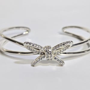 Puede incluir: Una pulsera de puño plateada con un diseño de lazo. El lazo está adornado con pequeños cristales brillantes. La pulsera tiene un diseño de doble banda y está sobre un fondo blanco.