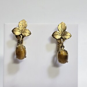 May include: A pair of vintage clip-on earrings. Each earring features a gold-toned leaf design at the top, with a dangling oval-shaped tiger's eye stone. The earrings are set against a white background.