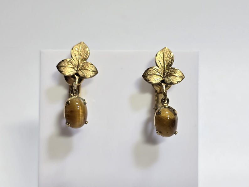 May include: A pair of vintage clip-on earrings. Each earring features a gold-toned leaf design at the top, with a dangling oval-shaped tiger's eye stone. The earrings are set against a white background.