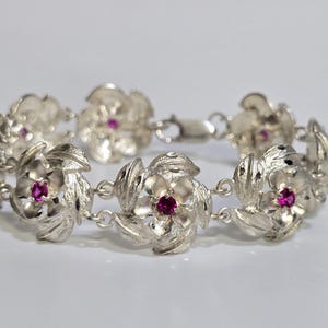 May include: A silver bracelet featuring floral designs with a small, round, red gemstone in the center of each flower. The bracelet is linked together with a clasp.