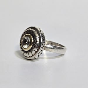 Puede incluir: Un anillo de plata con un engaste redondo y ornamentado con una gema transparente central. El engaste tiene un c&iacute;rculo interior dorado y patrones detallados y arremolinados. La banda es de plata pulida simple.