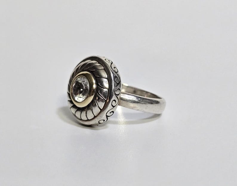 Puede incluir: Un anillo de plata con un engaste redondo y ornamentado con una gema transparente central. El engaste tiene un c&iacute;rculo interior dorado y patrones detallados y arremolinados. La banda es de plata pulida simple.