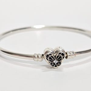 Könnte beinhalten: Ein silbernes Armkettchen mit einem herzförmigen Anhänger, der ein detailliertes Schmetterlingsdesign und einen kleinen, klaren Stein aufweist.