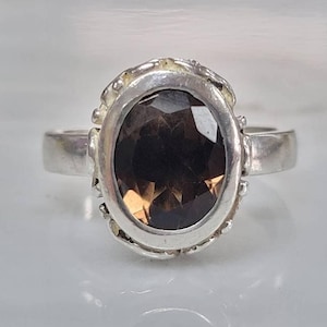 May include: A silver ring with an oval-shaped smoky quartz gemstone set in a textured bezel. The ring has a simple band.