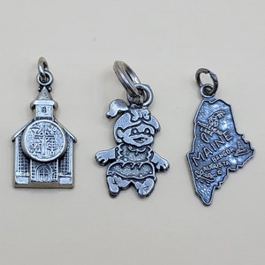May include: Three silver charm pendants. One is a church, one is a cartoon character, and one is the state of Maine.