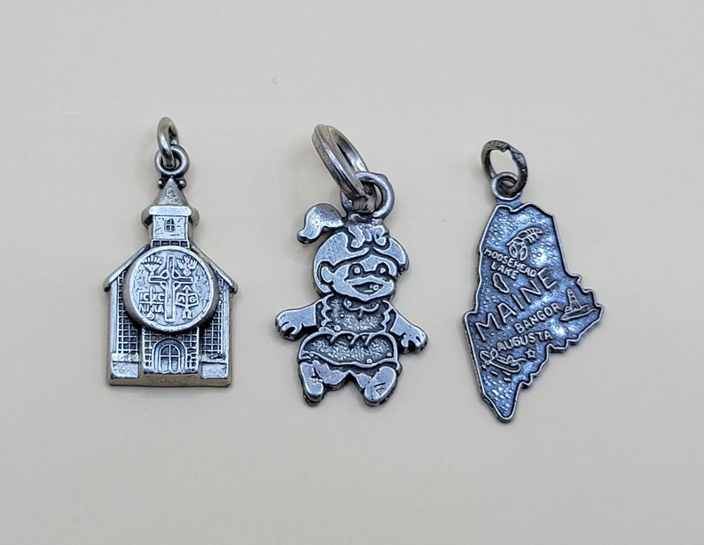 May include: Three silver charm pendants. One is a church, one is a cartoon character, and one is the state of Maine.