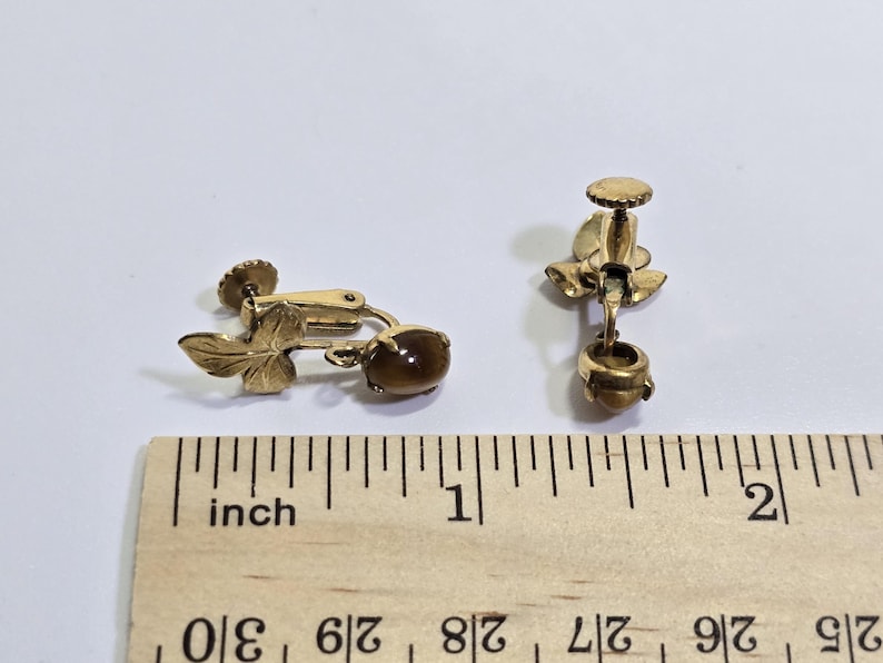 May include: A pair of vintage gold-toned clip-on earrings. Each earring features a leaf design and a brown, oval-shaped stone. The earrings are approximately 1 inch in length.