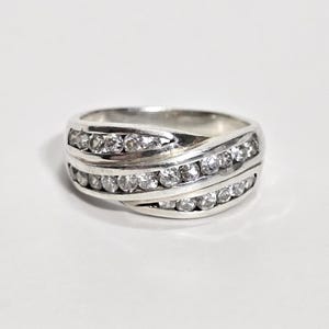 May include: A silver ring with a wave design, set with multiple clear, round-cut gemstones. The ring features three rows of stones, creating a sparkling, elegant look. The ring is on a white surface.