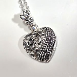 May include: A silver heart-shaped pendant necklace with an intricate floral design. The pendant has a textured surface and a delicate chain.