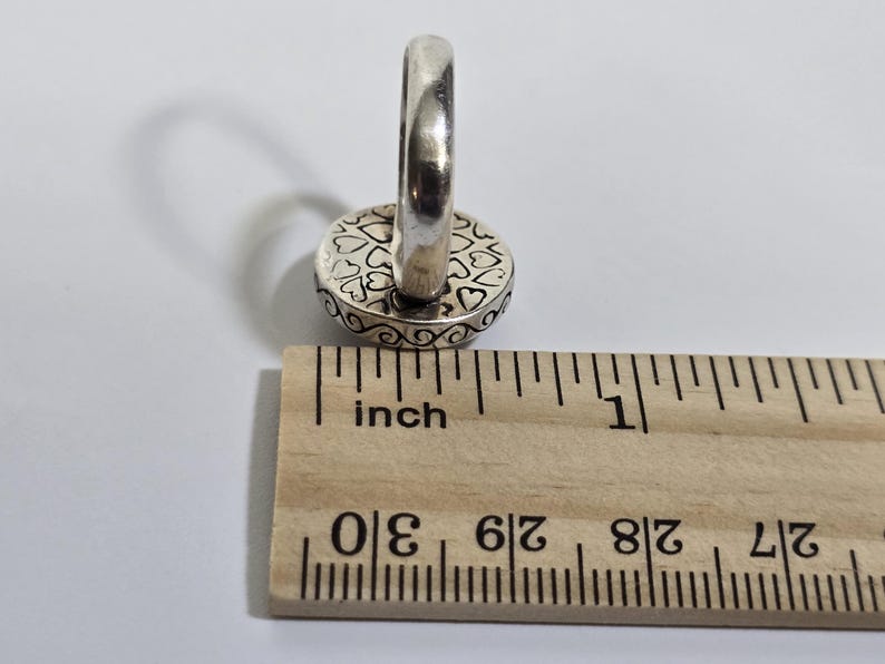 Puede incluir: Anillo de plata con una cara redonda con un dise&ntilde;o de coraz&oacute;n. El anillo est&aacute; junto a una regla que muestra medidas en pulgadas. La cara del anillo tiene aproximadamente 1,3 cm de di&aacute;metro.