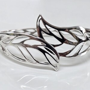 May include: A silver bangle bracelet with a leaf design. The bracelet is open and has a delicate, intricate design.