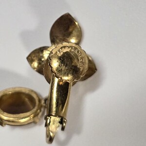 May include: A gold-toned vintage earring with a floral design. The earring features three petal-shaped elements and a small, round, open-backed setting. The earring's post and clasp are visible, indicating a clip-on style.