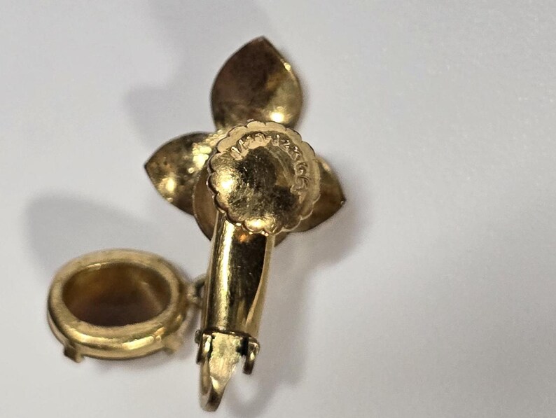 May include: A gold-toned vintage earring with a floral design. The earring features three petal-shaped elements and a small, round, open-backed setting. The earring's post and clasp are visible, indicating a clip-on style.