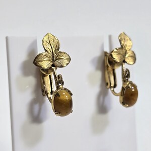 May include: A pair of vintage clip-on earrings. Each earring features a gold-toned, textured leaf design at the top, with a dangling oval-shaped, brown gemstone. The earrings are displayed against a white background.