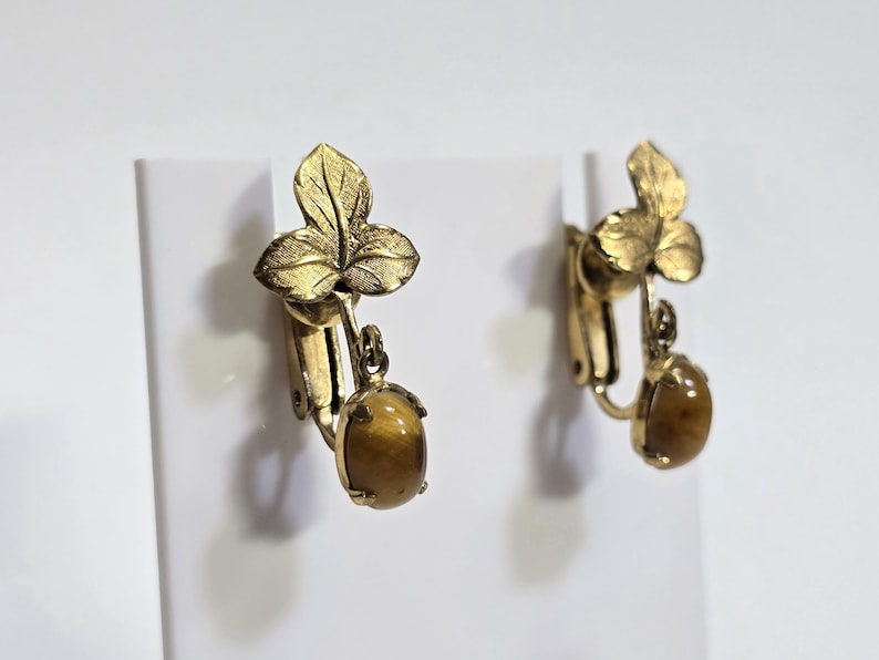 May include: A pair of vintage clip-on earrings. Each earring features a gold-toned, textured leaf design at the top, with a dangling oval-shaped, brown gemstone. The earrings are displayed against a white background.