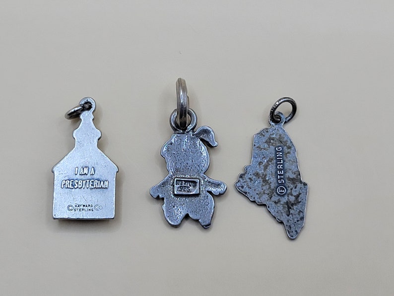 May include: Three silver charm pendants. The first is a church with the text "I am a Presbyterian" and "Dayware Co. Sterling". The second is a girl with the text "Mexico 925" on the back. The third is a state outline with the text "Sterling" on the back.