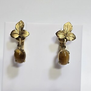 May include: A pair of gold-toned clip-on earrings. Each earring features a three-leaf design at the top and a dangling oval-shaped, amber-colored stone. The earrings are displayed against a white background.
