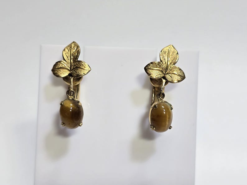 May include: A pair of gold-toned clip-on earrings. Each earring features a three-leaf design at the top and a dangling oval-shaped, amber-colored stone. The earrings are displayed against a white background.