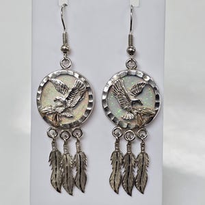 May include: Silver earrings with a white opal dreamcatcher design featuring an eagle in flight. The earrings have three silver feathers dangling from each dreamcatcher.